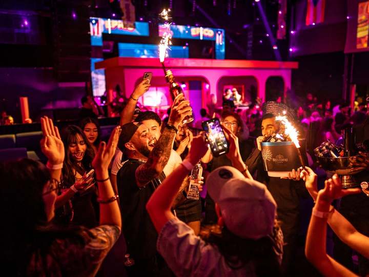 Público eufórico levantando botellas con bengalas frente al escenario rosa de una fiesta de reguetón de EPICO