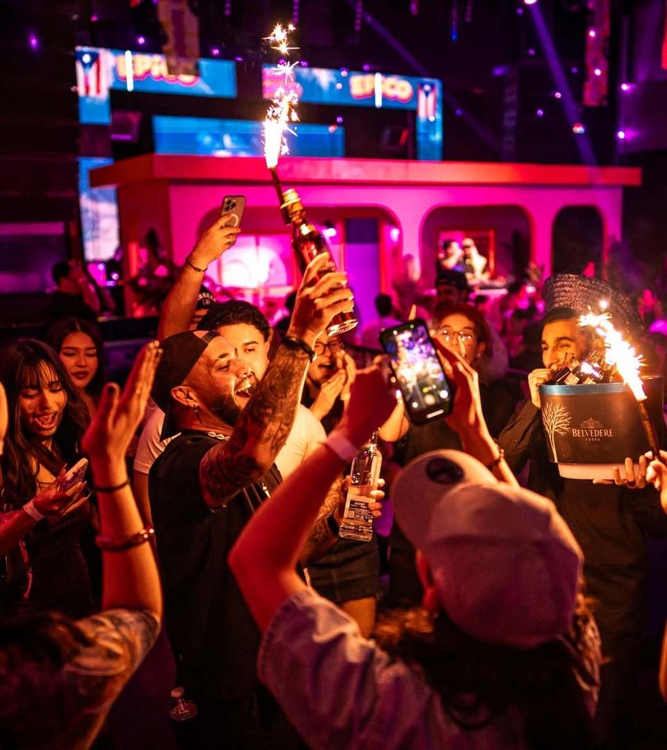 Cheering crowd raising bottles topped with sparklers in front of the pink stage at an EPICO reggaeton party