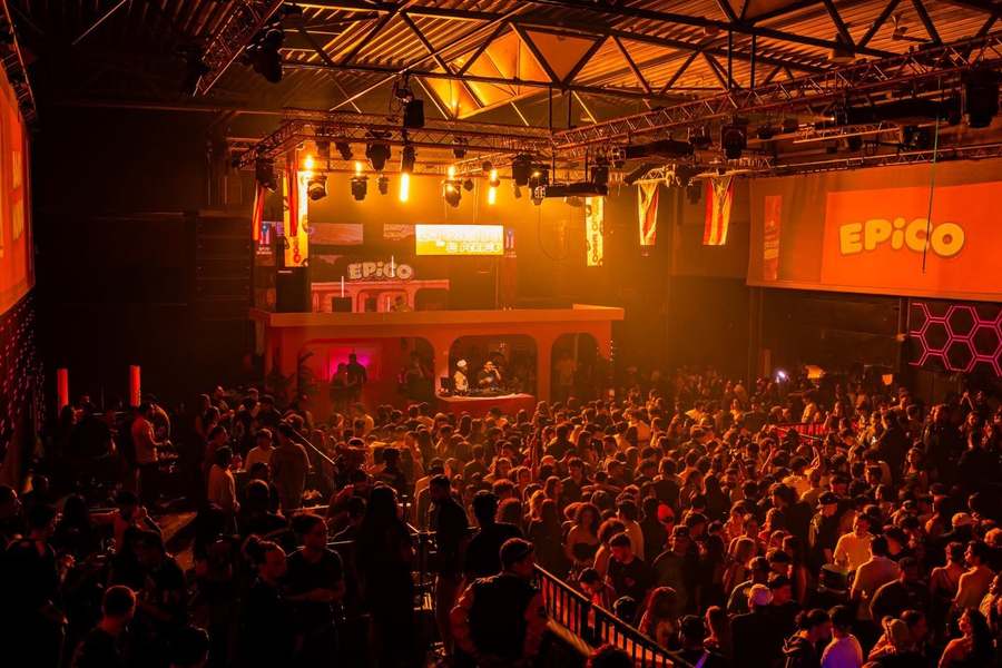 Sala llena bañada de luz naranja bajo los carteles de EPICO, vista desde el fondo de la fiesta